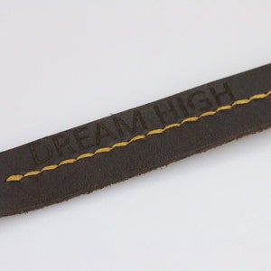 May include: Close-up of a dark brown leather strap with the words "DREAM HIGH" embossed in the leather. The strap has yellow stitching and a metal snap closure. The background is plain white.