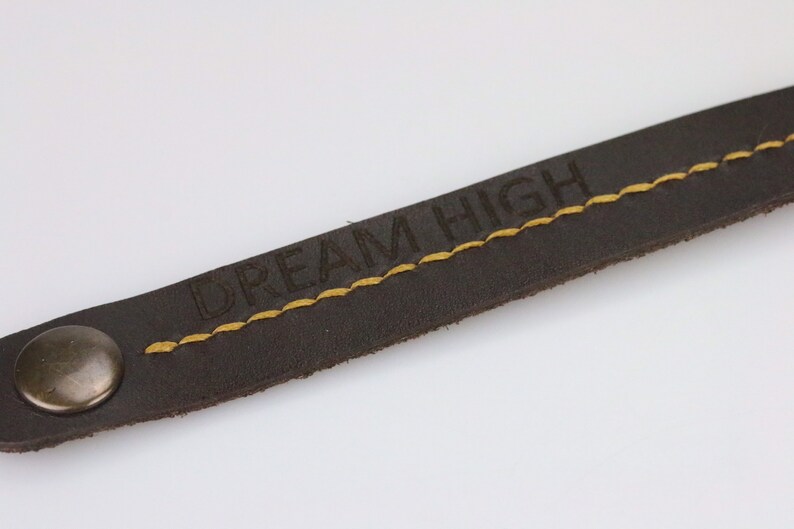 May include: Close-up of a dark brown leather strap with the words "DREAM HIGH" embossed in the leather. The strap has yellow stitching and a metal snap closure. The background is plain white.