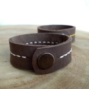 May include: Two brown leather bracelets with white stitching and a bronze button closure. The bracelets are circular and appear to be handmade. One bracelet is in focus, while the other is slightly blurred in the background. The bracelets are on a wooden surface.