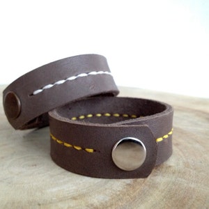 May include: Two brown leather bracelets with contrasting stitching. One bracelet has white stitching, and the other has yellow stitching. Each bracelet has a snap closure with a silver button.