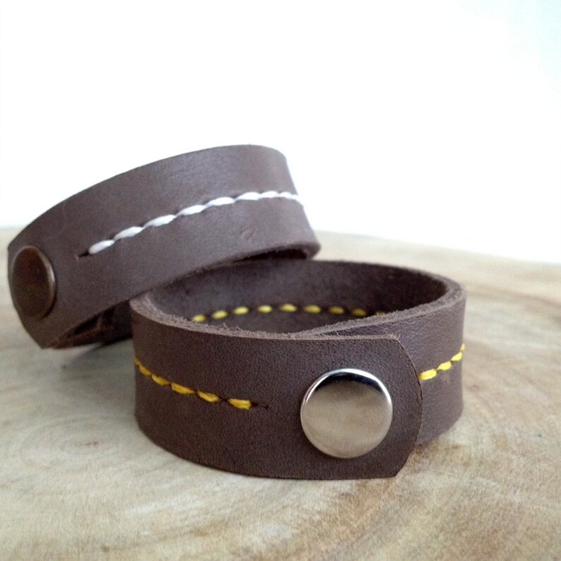 May include: Two brown leather bracelets with contrasting stitching. One bracelet has white stitching, and the other has yellow stitching. Each bracelet has a snap closure with a silver button.