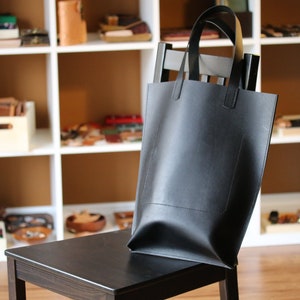 May include: A black leather tote bag with a rectangular shape and two handles. The bag is sitting on a black wooden chair. Shelves filled with leather goods are in the background.