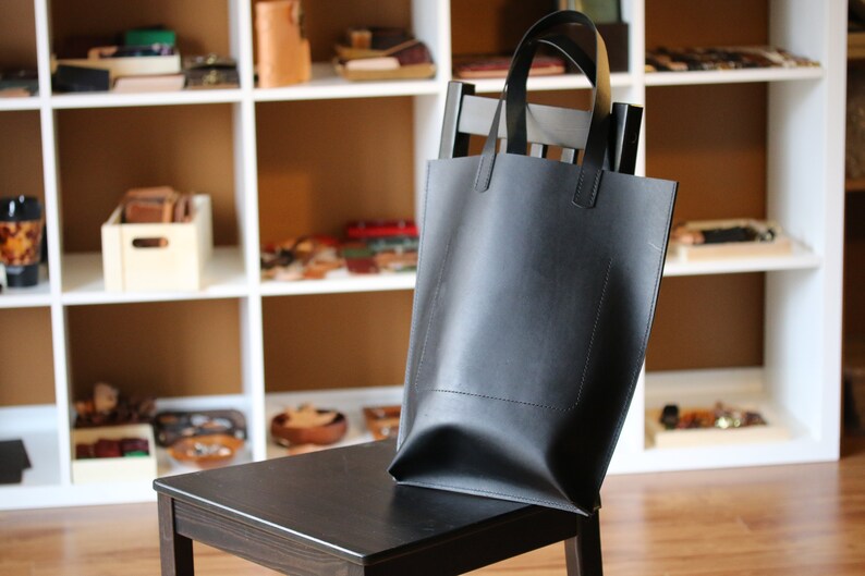 May include: A black leather tote bag with a rectangular shape and two handles. The bag is sitting on a black wooden chair. Shelves filled with leather goods are in the background.