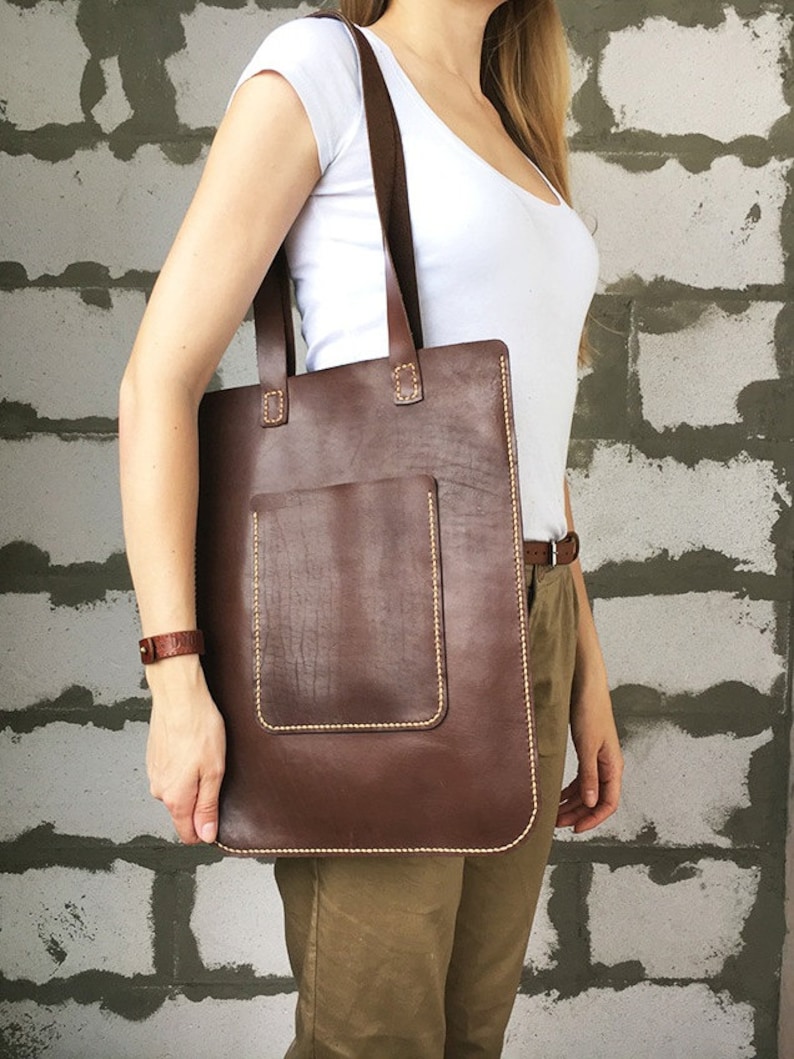 May include: A brown leather tote bag with a front pocket and a stitched design. The bag has two brown leather straps.