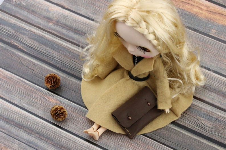 May include: A doll with blonde hair wearing a tan coat and holding a brown leather clutch. The doll is lying on a wooden surface, with two small pine cones nearby. The doll's coat has a black belt.