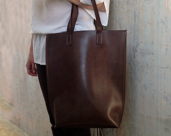 May include: A large, rectangular brown leather tote bag with two handles. The bag has visible stitching along the edges and at the handle attachments. The bag is held by a person, and the background is a textured gray wall.