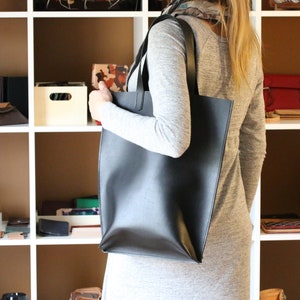 May include: A black leather tote bag with a shoulder strap is held by a person. The bag is rectangular and appears to be empty. Shelves in the background display various leather goods, including wallets and small bags.