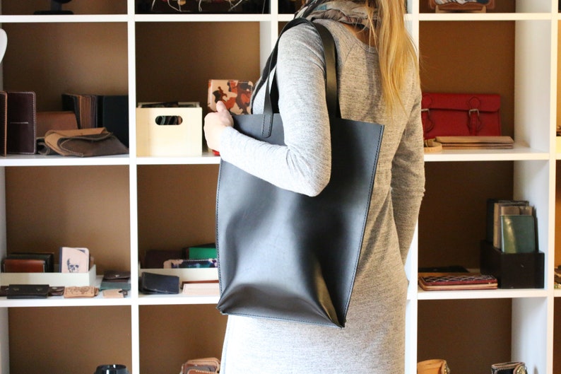May include: A black leather tote bag with a shoulder strap is held by a person. The bag is rectangular and appears to be empty. Shelves in the background display various leather goods, including wallets and small bags.