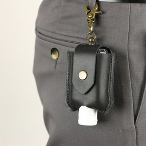 May include: A black leather hand sanitizer holder with a brass clip and a snap closure. The holder is attached to a belt loop.