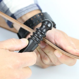 May include: A person using a black leather working tool to smooth out a piece of brown leather.