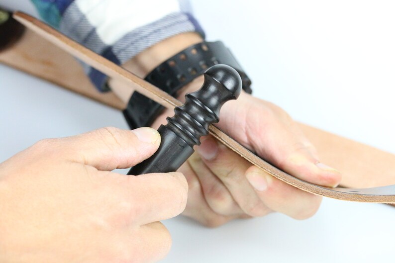 May include: A person using a black leather working tool to smooth out a piece of brown leather.