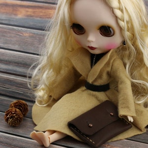May include: A Blythe doll with long blonde hair, wearing a tan coat and a black belt. The doll is holding a small brown leather wallet. Two small pinecones are next to the doll on a wooden surface.