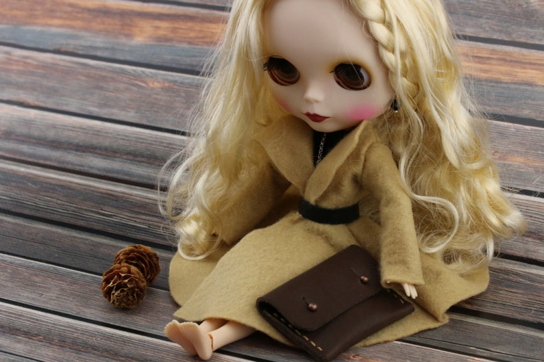 May include: A Blythe doll with long blonde hair, wearing a tan coat and a black belt. The doll is holding a small brown leather wallet. Two small pinecones are next to the doll on a wooden surface.
