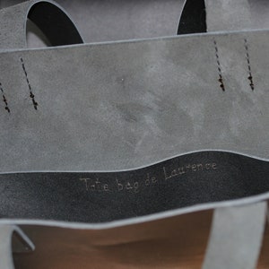 May include: Close-up of a gray tote bag with a black interior. The bag has two straps and the text "Tote bag de Laurence" is visible inside. The bag is made of a soft material.