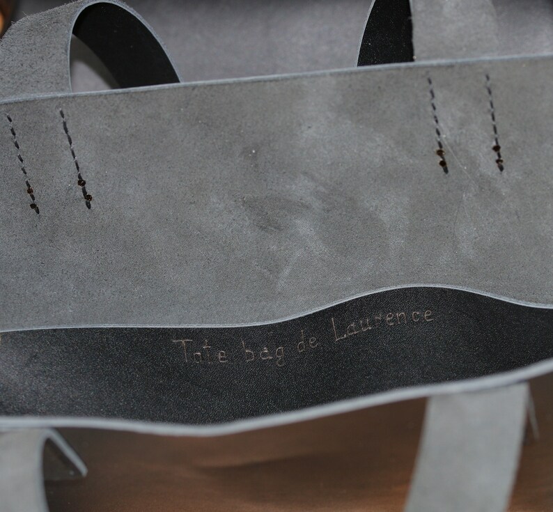 May include: Close-up of a gray tote bag with a black interior. The bag has two straps and the text "Tote bag de Laurence" is visible inside. The bag is made of a soft material.