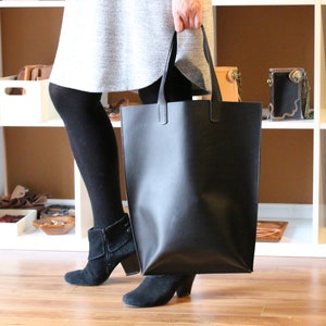 May include: A large, black leather tote bag with two handles. The bag is rectangular and appears to be empty. The person holding the bag is wearing black tights and black suede ankle boots. The background includes white shelves with various leather goods.