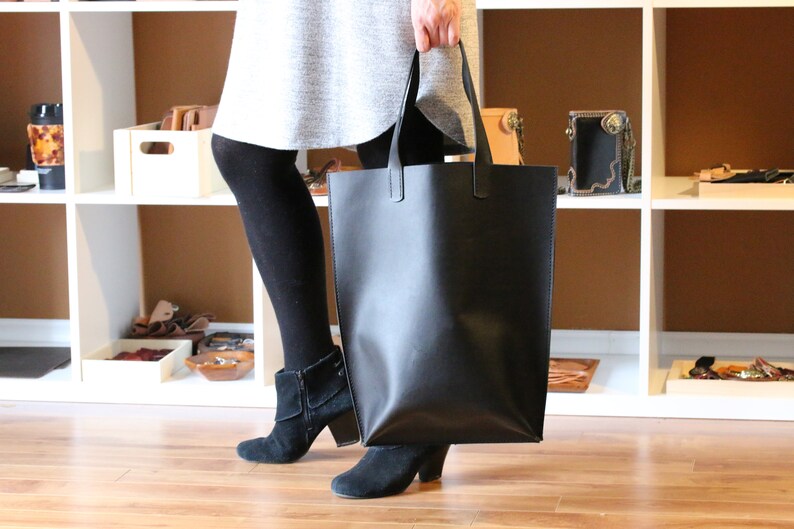 May include: A large, black leather tote bag with two handles. The bag is rectangular and appears to be empty. The person holding the bag is wearing black tights and black suede ankle boots. The background includes white shelves with various leather goods.