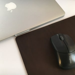 May include: A black wireless mouse with the word "RAPOO" printed on the side, sitting on a brown leather mouse mat next to a silver laptop.