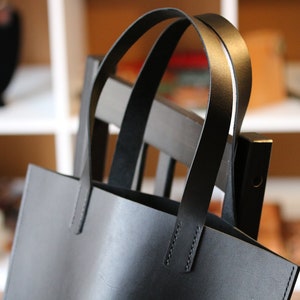 May include: A black leather tote bag with two arched handles. The bag is rectangular with a simple design and visible stitching. The bag is hanging on a dark wooden chair. The background is blurred, showing shelves and other items.