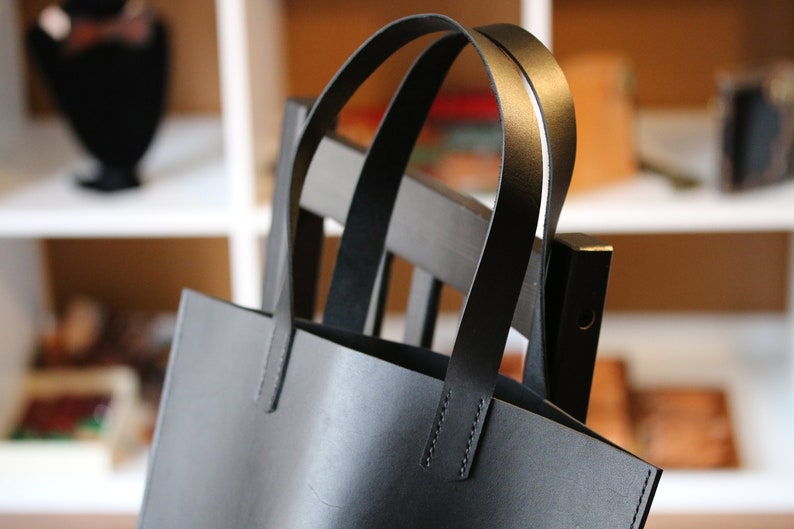May include: A black leather tote bag with two arched handles. The bag is rectangular with a simple design and visible stitching. The bag is hanging on a dark wooden chair. The background is blurred, showing shelves and other items.