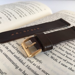 May include: Two brown leather watch straps with gold buckles. The straps are lying on a book.
