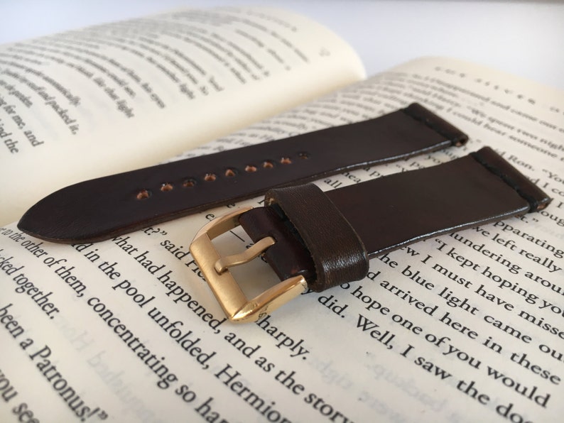May include: Two brown leather watch straps with gold buckles. The straps are lying on a book.