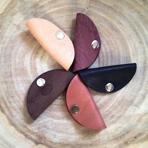 May include: Five leather keychains in the shape of a taco shell. The keychains are in different colors: brown, black, tan, and two shades of red. Each keychain has a silver metal snap closure.