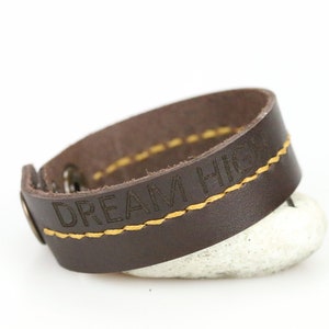 Könnte beinhalten: Ein braunes Lederarmband mit den Worten "DREAM HIGH" auf der Vorderseite. Das Armband hat gelbe Nähte und einen Metallverschluss. Das Armband liegt auf einem weißen Stein.