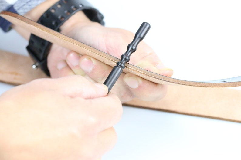 May include: A person using a black leather crafting tool to punch holes in a piece of brown leather.