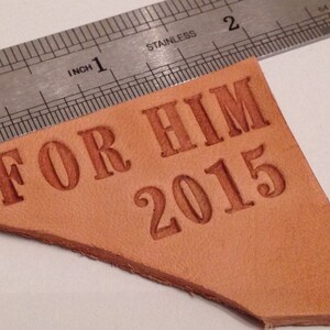 K&ouml;nnte beinhalten: Ein braunes Lederst&uuml;ck mit den Worten "FOR HIM 2015" in brauner Pr&auml;gung.