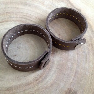 May include: Two brown leather bracelets with white stitching and snap closures. The bracelets are circular and have a simple, rustic design. The stitching is visible along the edges, adding a handcrafted touch.
