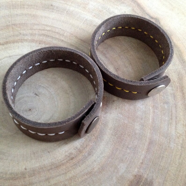 May include: Two brown leather bracelets with white stitching and snap closures. The bracelets are circular and have a simple, rustic design. The stitching is visible along the edges, adding a handcrafted touch.