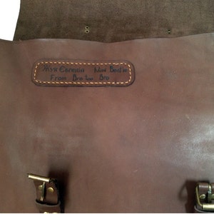 May include: Brown leather messenger bag with a personalized inscription that reads "Anya Eremenia Mimi Beattie From Bro Two Bro". The bag has two buckles on the front.