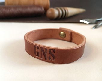 May include: A brown leather bracelet with the initials "GNS" engraved on the front. The bracelet has a simple, minimalist design with a snap closure. The leather appears to be of high quality, with a natural grain and texture. The bracelet is approximately 1 inch wide.