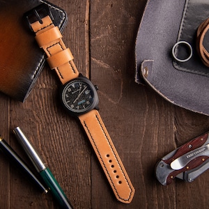 May include: A black watch with a tan leather strap, a black pen, a green pen, a brown leather wallet, a grey leather pouch with a brown leather strap, and a silver pocket knife with a wooden handle.
