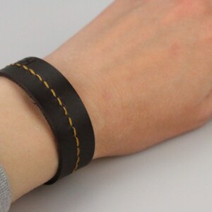 May include: A dark brown leather bracelet with yellow stitching is worn on a wrist. The bracelet is a simple band design, with two parallel lines of yellow stitching. The leather appears to be smooth and the bracelet is a classic accessory.