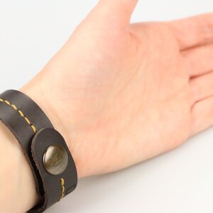 May include: Dark brown leather bracelet with yellow stitching and a metal button clasp. The bracelet is wrapped around a wrist, showcasing the design and craftsmanship of the accessory. A simple, yet stylish piece.