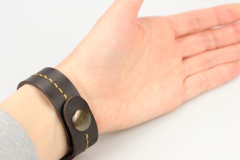 May include: Dark brown leather bracelet with yellow stitching and a metal button clasp. The bracelet is wrapped around a wrist, showcasing the design and craftsmanship of the accessory. A simple, yet stylish piece.