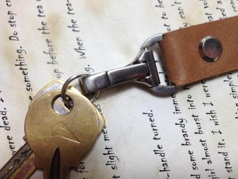 Puede incluir: Primer plano de un llavero de cuero marr&oacute;n con un cierre met&aacute;lico y varias llaves. Las llaves est&aacute;n unidas a un anillo met&aacute;lico. El fondo es un papel vintage con texto manuscrito.
