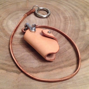 K&ouml;nnte beinhalten: Ein hellbrauner Leder-Schl&uuml;sselanh&auml;nger mit einem kleinen Lederbeutel. Der Beutel hat einen Metall-Druckknopfverschluss und einen Metallring zum Anbringen von Schl&uuml;sseln.