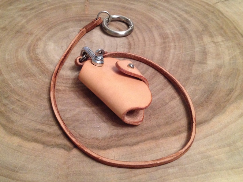 K&ouml;nnte beinhalten: Ein hellbrauner Leder-Schl&uuml;sselanh&auml;nger mit einem kleinen Lederbeutel. Der Beutel hat einen Metall-Druckknopfverschluss und einen Metallring zum Anbringen von Schl&uuml;sseln.