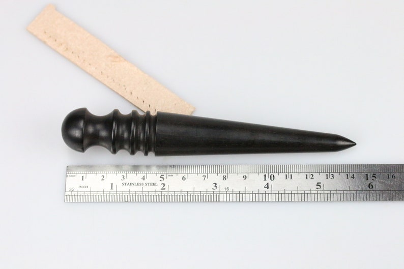 May include: A black wooden leather stitching tool with a rounded handle and a pointed tip. The tool is measured against a ruler that shows it is 6 inches long.