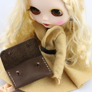 May include: A brown leather doll-sized wallet with copper rivets and white stitching. The wallet is open, revealing the interior. The doll is wearing a tan coat with a black belt and has long blonde hair.