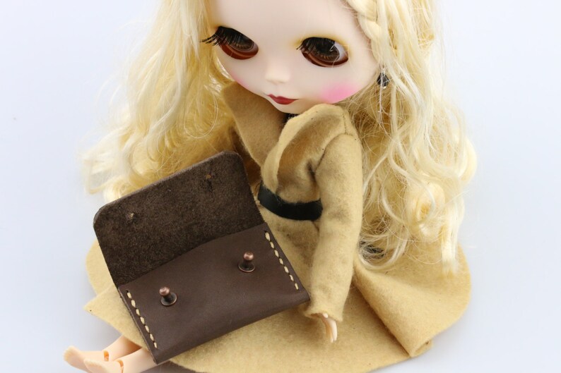 May include: A brown leather doll-sized wallet with copper rivets and white stitching. The wallet is open, revealing the interior. The doll is wearing a tan coat with a black belt and has long blonde hair.