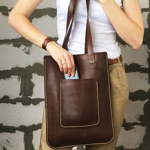May include: A brown leather tote bag with a large front pocket and a stitched detail. The bag has two long straps.