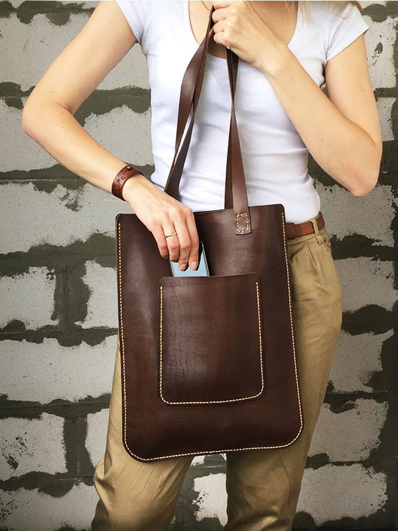 May include: A brown leather tote bag with a large front pocket and a stitched detail. The bag has two long straps.