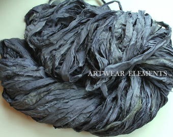 May include: A bundle of dark charcoal gray silk ribbon with a crinkled, textured finish. The fabric strips are loosely coiled together in a heap. The text ARTWEAR ELEMENTS is printed in white on the right side of the ribbon bundle.
