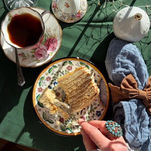 Può includere: Una tavola imbandita con una fetta di torta a strati su un piatto decorativo, una tazza di tè e un vaso decorativo. Il piatto e la tazza hanno disegni floreali. Una mano con un anello di turchese e pietra rossa tiene una forchetta.