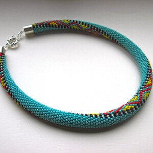May include: A turquoise beaded necklace with a silver clasp. The necklace features a pattern of red, yellow, black, and white beads. The necklace is a circular shape and is likely worn around the neck.