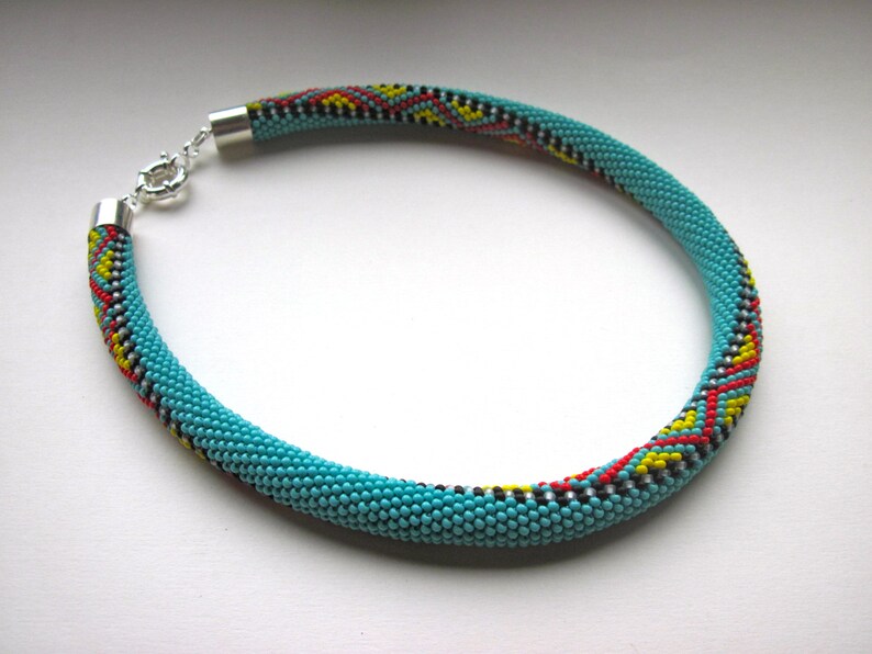 May include: A turquoise beaded necklace with a silver clasp. The necklace features a pattern of red, yellow, black, and white beads. The necklace is a circular shape and is likely worn around the neck.